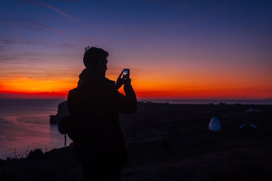 Man traveler silhouette hiking at sunset on Santorini island. Tourist taking photo of night landscape on smartphone