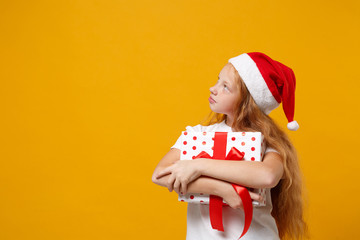 Pensive little ginger kid Santa girl 12-13 years old in white t-shirt Christmas hat isolated on yellow background. New Year 2020 celebration concept. Mock up copy space. Hold box with gift ribbon bow.