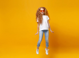 Pretty little ginger kid girl 12-13 years old in white t-shirt, heart glasses isolated on yellow wall background. Childhood lifestyle concept. Mock up copy space. Having fun, fooling around, jumping.
