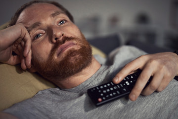Portrait of bearded adult man watching TV at night while lying on couh in dark room and switching...