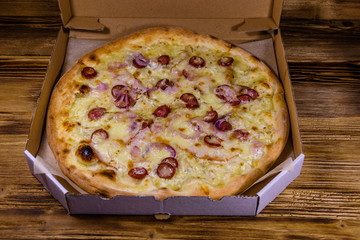 Pizza with sausages, ham and parmesan cheese in cardboard box on a wooden table