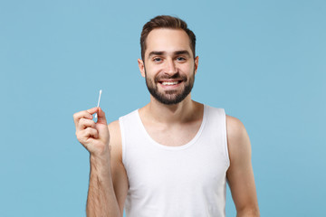 Bearded man 20s years old in white shirt hold cotton swab stick for ear cleaning isolated on blue...