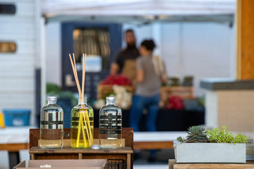 Attractive display of clear bottles of liquid aromatherapy with reed diffusers at an outdoor market with defocused shopper and vendor in the background.