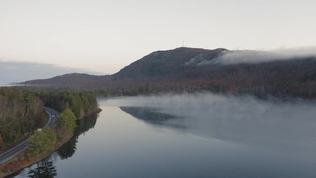 Two vehicles driving around a curve on the shore of a misty pond with a mountain in the distance AERIAL SLIDE