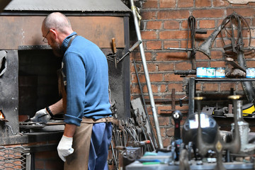 Fototapeta premium An experienced blacksmith works with open fire. A blacksmith extinguishes a flame in a forge with a spark firework, forges hot iron in a workshop