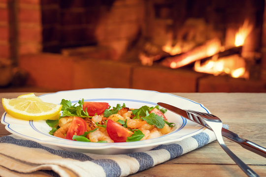 Fresh homemade salad of shrimp, arugula and tomato in a white plate on a wooden table in a room with a fireplace, close-up