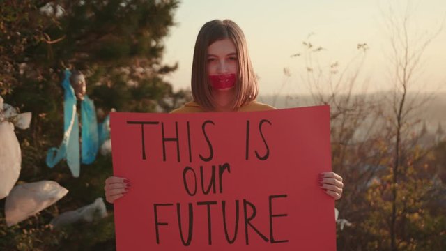 Young woman activist with mouth glued holding encouraging red poster This is our future standing at landfill site with garbage nature environmental community earth eco outdoor recycle trash waste