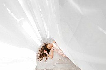 Portrait from above of little beautiful girl with long hair lying on floor with white curtain  covering her