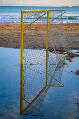Football goal reflected from the water in a puddle. The beach soccer field is flooded with water after the storm. Football goal against the backdrop of the sea and sunset sky. Roses, Catalonia, Spain.
