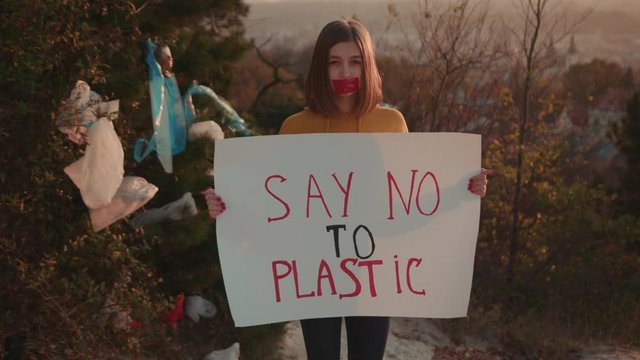 Young woman activist with glued mouth holding encouraging white poster Say no to plastic standing at landfill site with garbage nature environmental community earth eco environment outdoor slow motion