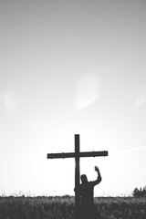 Vertical shot of a male standing near a cross with his hand up in black and white