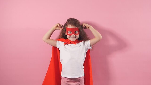 Beautiful little curly girl wearing red hero suit and mask showing how she is strong isolated over pink background