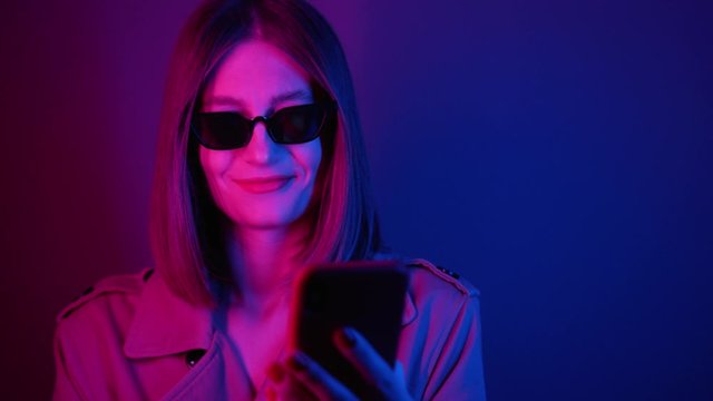 Shooting of charming young woman in stylish trench and sunglasses using red smartphone. Surprised girl reading message on screen. Technology, news, neon lights, beauty, purple