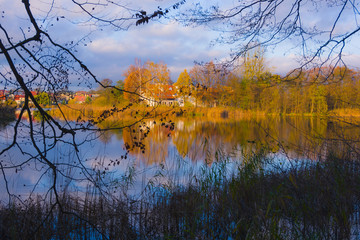 Colorful autumn landscape, reflection of yellow and red leaves of trees and white building in the lake, shore covered with reeds, view through branches