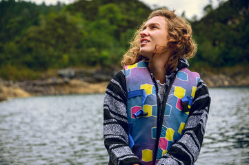 Fototapeta premium Young woman sitting on a wooden raft in the middle of a turqeuza blue lake admiring the landscape of wooded mountains on a cloudy day