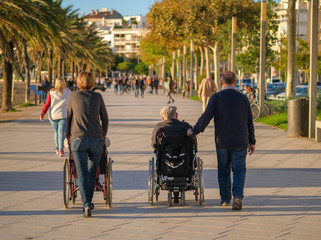 Adult children spend time with their parents with disabilities in wheelchairs. Family walks along the promenade on a sunny day. A wheelchair is being pushed by the accompanying people. Roses Catalonia