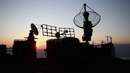 Creative artwork decoration. Silhouette of mobile air defence truck with radar antenna during sunset. Satellite dishes or radio antennas against evening sky. Selective focus