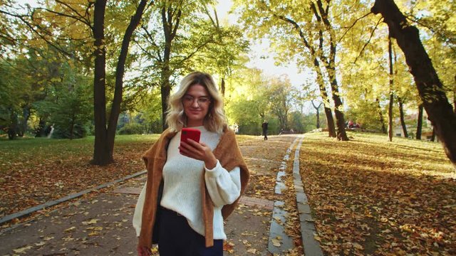 Attractive blonde lady using smartphone walking in beautiful park at fall season. Cheerful urban girl reading good news on mobile phone staying outside in nature.