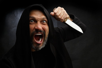 Portrait of a bald man with a beard in a black hood with a knife on a dirty gray background. Maniac.