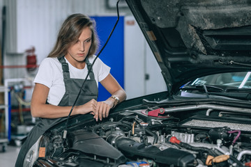 Photo of young female car repair worker.Beautiful brunette wearing black jumpsuit. Girl staying near the car in car servise working under car cowl.