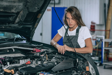 Fototapeta na wymiar Photo of young girl car repair worker.Beautiful brunette wearing black jumpsuit. Girl staying near the car in car servise working under car cowl.