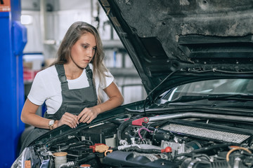 Young female from car service in a black jumpsuit and white t-shirt standing near the broken car. The girl opened the hood and looked at the engine. Refit woman car