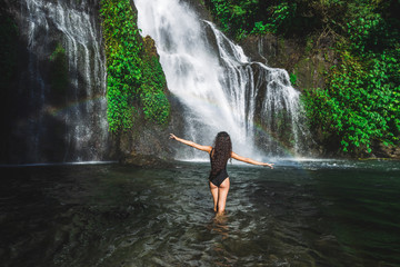 Young slim brunette woman with curly hair enjoying in lagoon of huge tropical waterfall Banyumala in Bali. Wearing in black swimsuit. Happy vacations in Indonesia. Wanderlust travel concept.