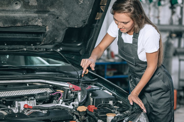 A beautiful girl in a black jumpsuit and a white t-shirt is smiling, checking the oil level in a black car in the garage.