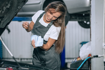 A beautiful girl in a black jumpsuit and a white t-shirt is smiling, checking the oil level in a black car in the garage.