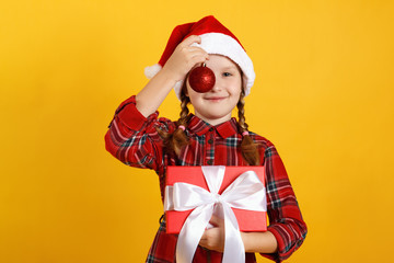 Fototapeta na wymiar Happy little girl with christmas ball toy and box with a gift. Close-up of a child in a santa hat and red dress on a yellow background
