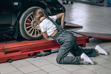 A beautiful girl in a white T-shirt and black jumpsuit is sitting near a black car removing the...