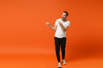 Fototapeta na wymiar Cheerful young man in casual white t-shirt posing isolated on bright orange wall background studio portrait. People sincere emotions lifestyle concept. Mock up copy space. Point index fingers aside.
