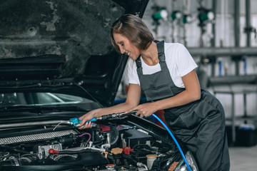 Obraz na płótnie Canvas A beautiful girl in a black jumpsuit and a white t-shirt is smiling, checking the oil level in a black car in the garage.