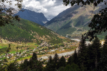 Mestia town below agriculture fields and big mountains seen from a trail on a sunny day with big clouds