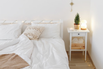 Little christmas tree in a pot and night salt lamp on bedside table. New year winter home interior decor. Christmas holiday decorations with led garland lights. White stylish cozy scandinavian bedroom