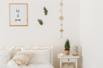 Little christmas tree in a pot on bedside table. New year winter home interior decor. Christmas holiday decorations. White stylish cozy bedroom: bed, led garland lights, frame with text LET IT SNOW.