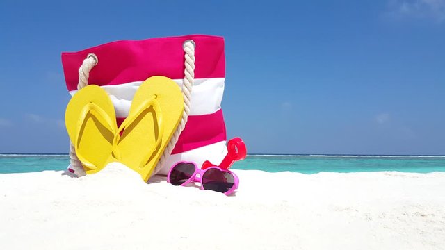 Summer set bag with yellow flip flops, pink sunglasses and beach toys over white sand of exotic beach on light blue sky background, concept design
