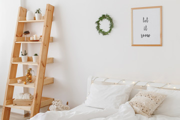 New year winter home interior decor. Simple Christmas holiday decorations: wreath of pine branches, led garland lights, frame with text LET IT SNOW. White stylish cozy scandinavian bedroom.