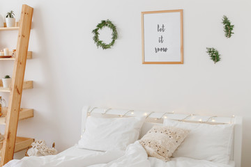 New year winter home interior decor. Simple Christmas holiday decorations: wreath, pine branches, coniferous twigs, led lights, frame with text LET IT SNOW. White stylish cozy scandinavian bedroom.