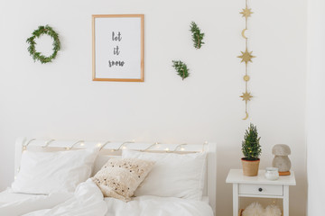 New year winter home interior decor. Simple Christmas holiday decorations: little christmas tree in a pot, wreath, pine branches, frame with text LET IT SNOW. White stylish cozy scandinavian bedroom.