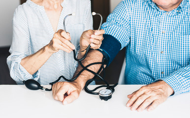 The therapist male hands in blue shirt with a classic tonometer measuring blood pressure of a man in home room on white table.