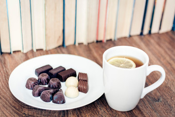 chocolate candies on white plate  and cup of tea with lemon with Stack of books on the background .