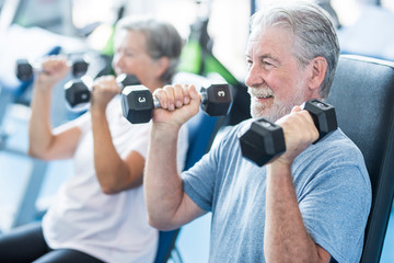mature man with his wife doing exercise of biceps at the gym - fitness and healthy lifestyle concept - training their body together - couple of two seniors - obrazy, fototapety, plakaty