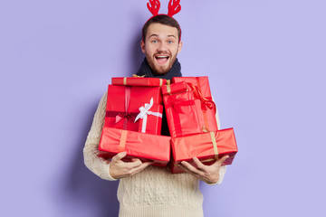 Cheerful glad male with various red christmas boxes in hands, happily yelling, satisfied by making presents to close friends, indoor studio shot, isolated photo