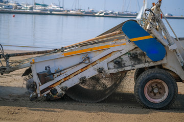 A special machine filters sand and removes waste and seaweed from it on blue sea background. Beach garbage cleaning machine at work.  Roses, Catalonia, Spain.