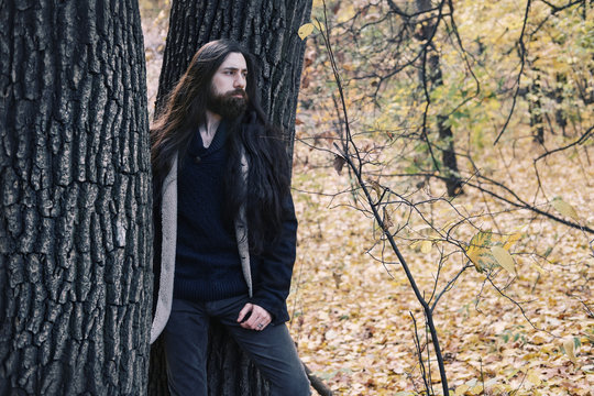 atmospheric autumn outdoor portrait of young man wearing long hair, beard and moustache, standing between trees