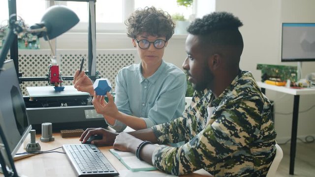 Woman is holding 3d printer model and talking to young African American man using computer in office, machine is printing plastic element in background.