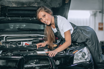 Fototapeta na wymiar A brunette in a black jumpsuit and a white t-shirt near the open hood. Young female in the garage is smiling at the camera.car repair concept