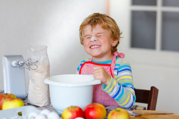 Cute little happy blond preschool kid boy baking apple cake and muffins in domestic kitchen. Funny...