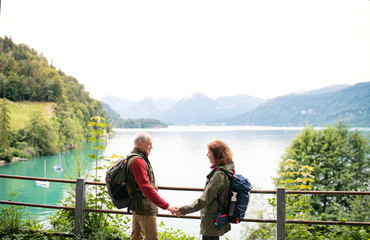 A senior pensioner couple hikers standing by lake in nature, talking.